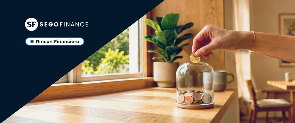 Mano introduciendo una moneda en un tarro de cristal con ahorros sobre una mesa de madera junto a una ventana